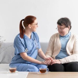 A health and social care giver in a blue uniform sitting with an elderly woman, holding hands and smiling warmly, with cups of tea on the table.
