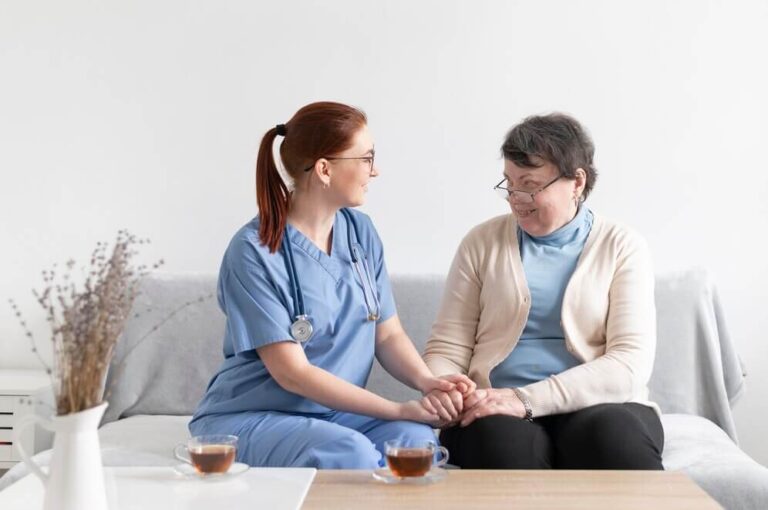 A health and social care giver in a blue uniform sitting with an elderly woman, holding hands and smiling warmly, with cups of tea on the table.