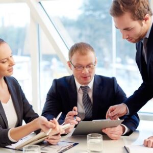 Three business professionals in a modern office discussing strategy and analyzing information on a tablet during a meeting of Executive Management.