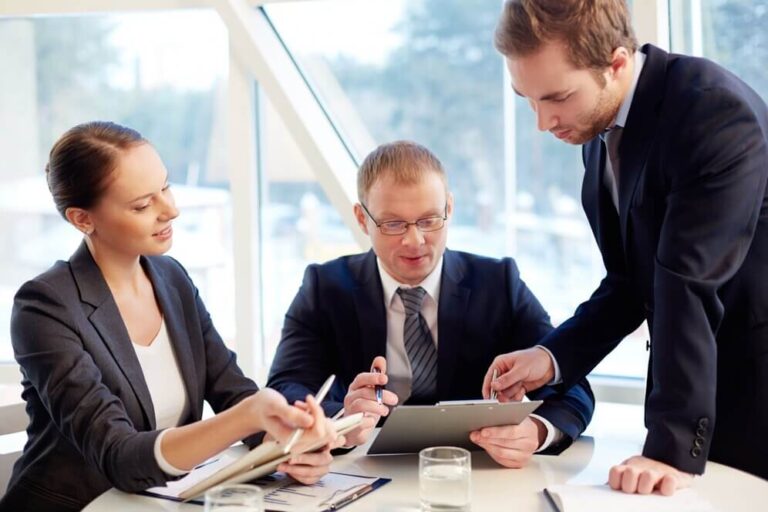 Three business professionals in a modern office discussing strategy and analyzing information on a tablet during a meeting of Executive Management.