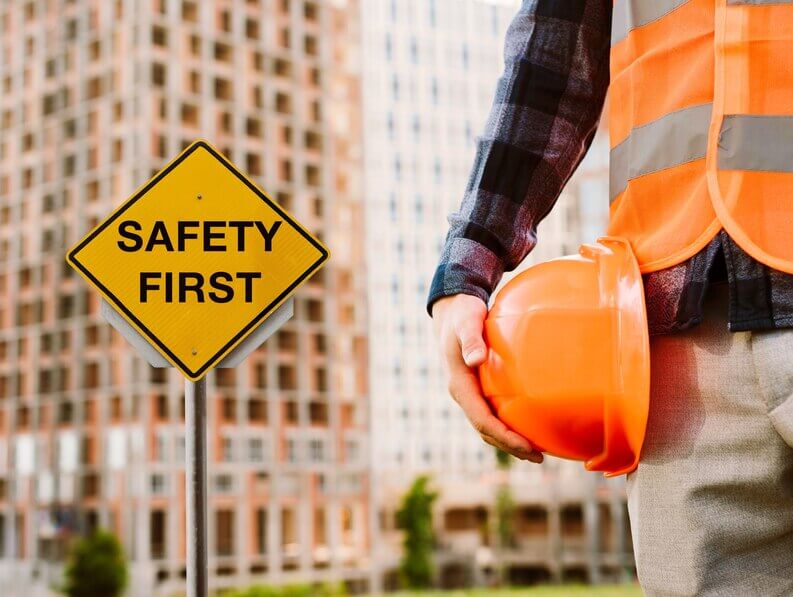 "Safety Orientation Course: Construction worker holding an orange safety helmet near a 'Safety First' sign in an urban setting."