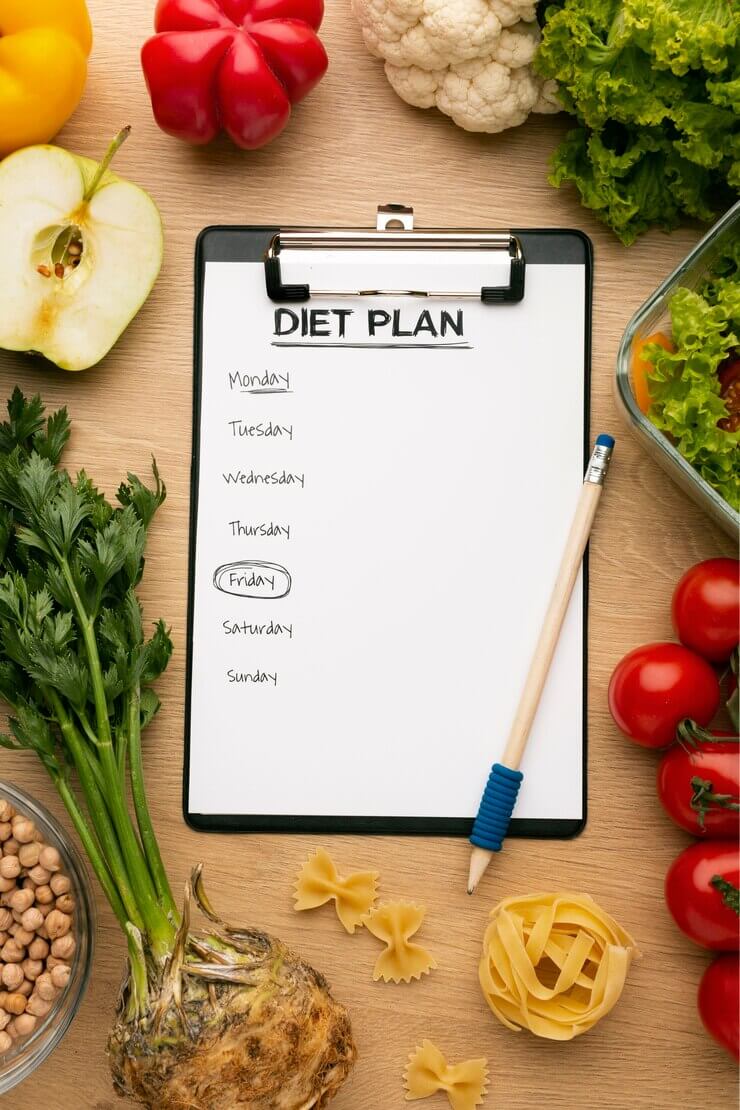 Clipboard with a weekly diet plan surrounded by fresh vegetables, fruits, pasta, and legumes on a wooden table.