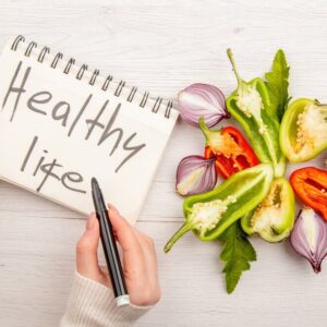 "Notebook with 'Healthy Life' written on it, placed next to colorful sliced vegetables like peppers and onions on a wooden table."