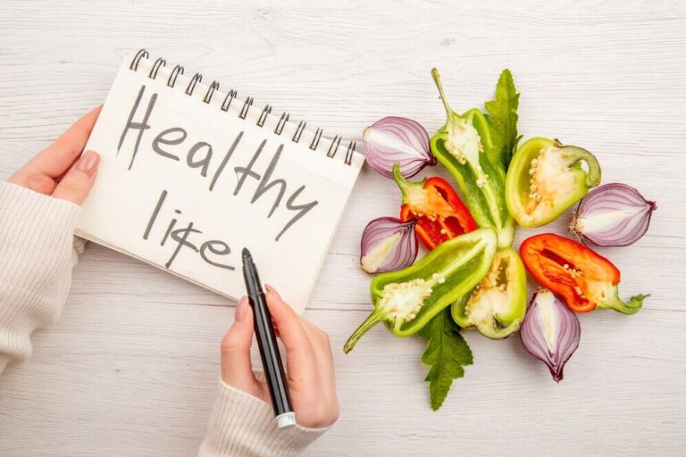 "Notebook with 'Healthy Life' written on it, placed next to colorful sliced vegetables like peppers and onions on a wooden table."