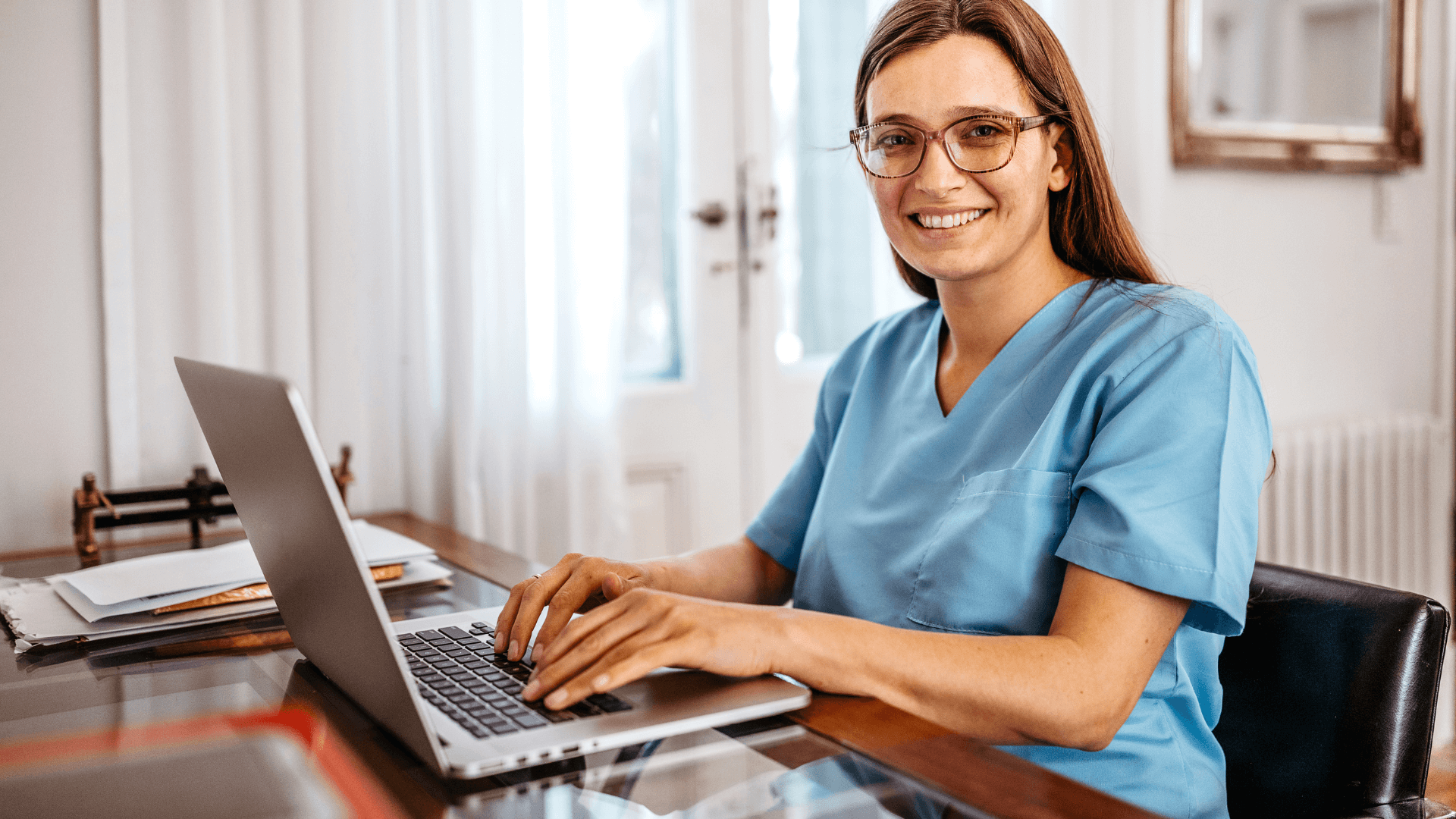 A smiling healthcare professional wearing blue scrubs sits at a desk, working on a laptop. The setting appears to be a well-lit home or office environment, with documents and office accessories on the table.